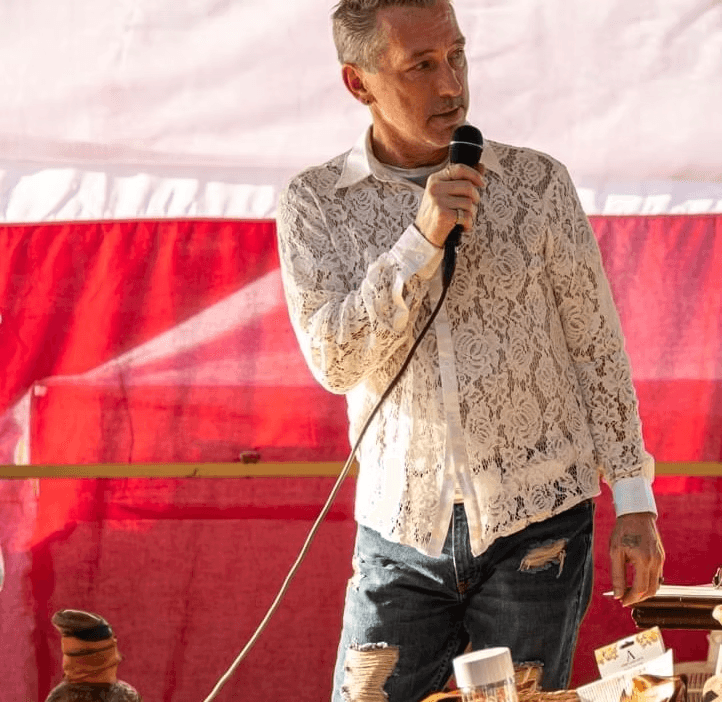 George I. Fricke III on the mic in a lace shirt at an outdoor event