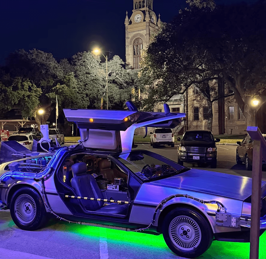 A DeLorean with gullwing doors up, lit green, parked by the Llano courthouse at night